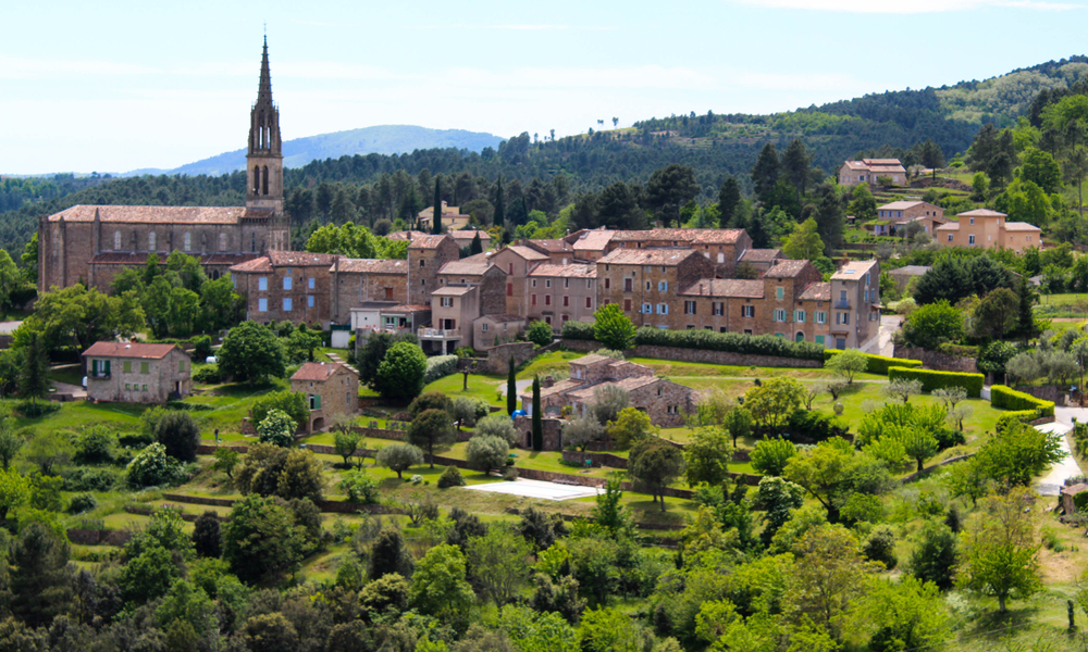 Stenen huisjes en een spitse klokkentoren gelegen op een groen stuk land en omringd door bergen, heggen en bomen.