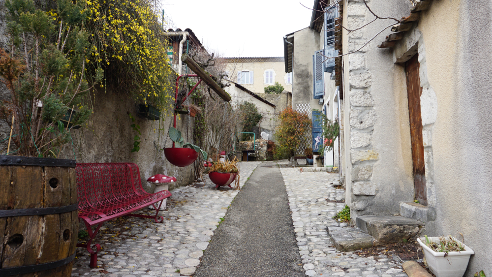 smal straatje met links een met struik begroeide muur en een rood bankje en rechts huizen