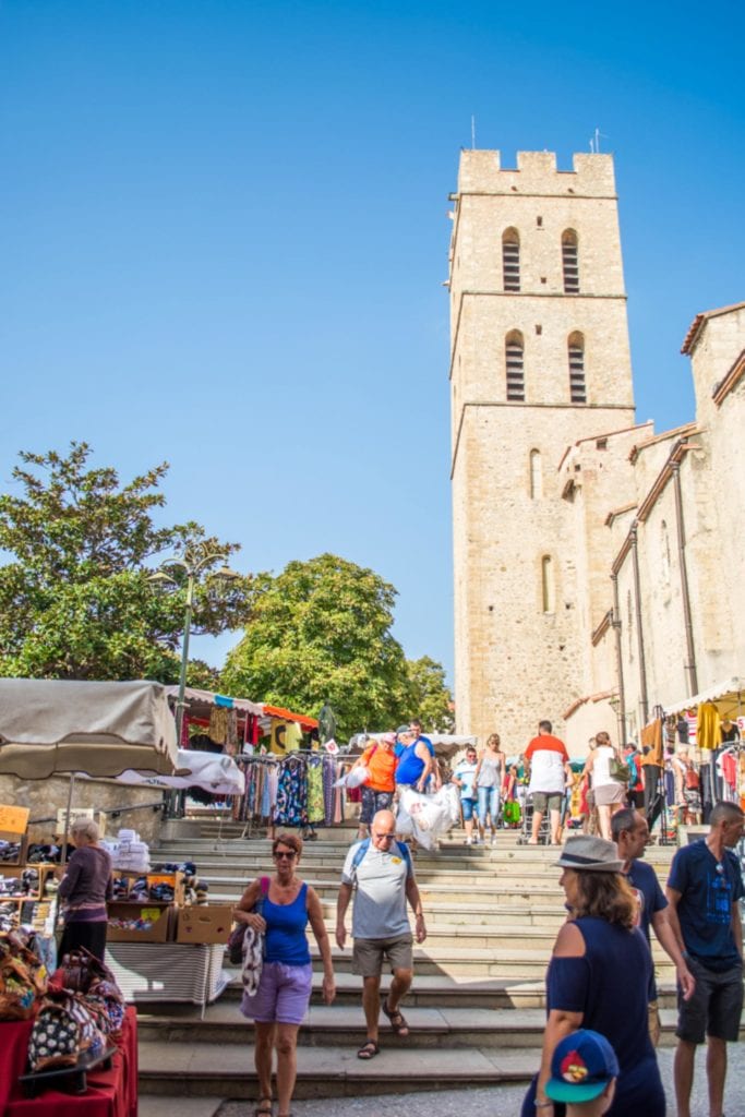 drukte tijdens de markt op een zomerse dag in het centrum van Argelès-sur-Mer. Er lopen zomers geklede mensen op de trap naast een kerkgebouw.